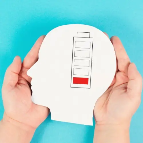 A pair of hands holding a white cutout of a human head with a low battery icon inside, symbolizing mental exhaustion and burnout on a bright blue background.