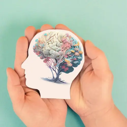 A pair of hands gently cupping a white cutout of a human head, which features a vibrant, flowering tree growing inside the brain area against a soft teal background.